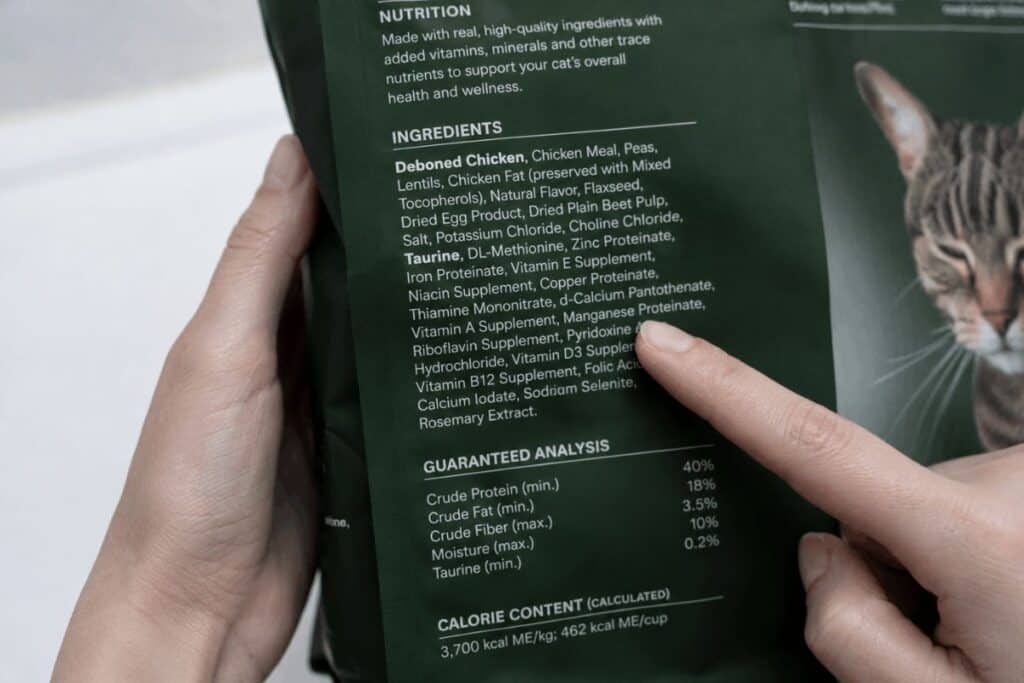 A person reading the ingredient label on a grain free cat food bag, with finger pointing to deboned chicken as the first ingredient and taurine listed below.